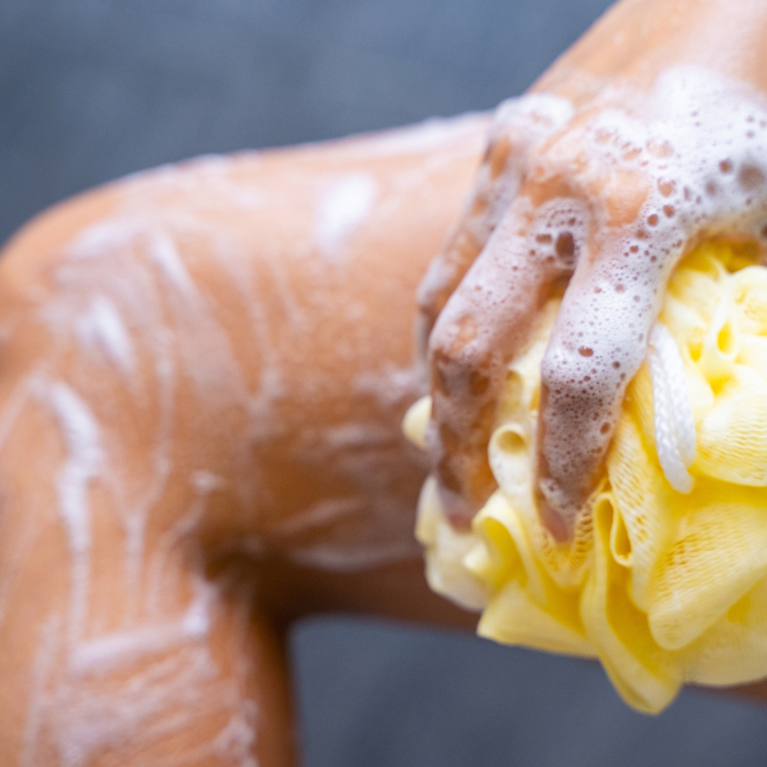 Person using vegan marshmallow body wash with lather and yellow sponge, highlighting soothing and moisturizing effects.