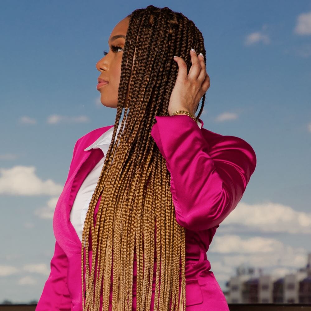 Woman with long braided hair standing outdoors against a blue sky, wearing a pink blazer.