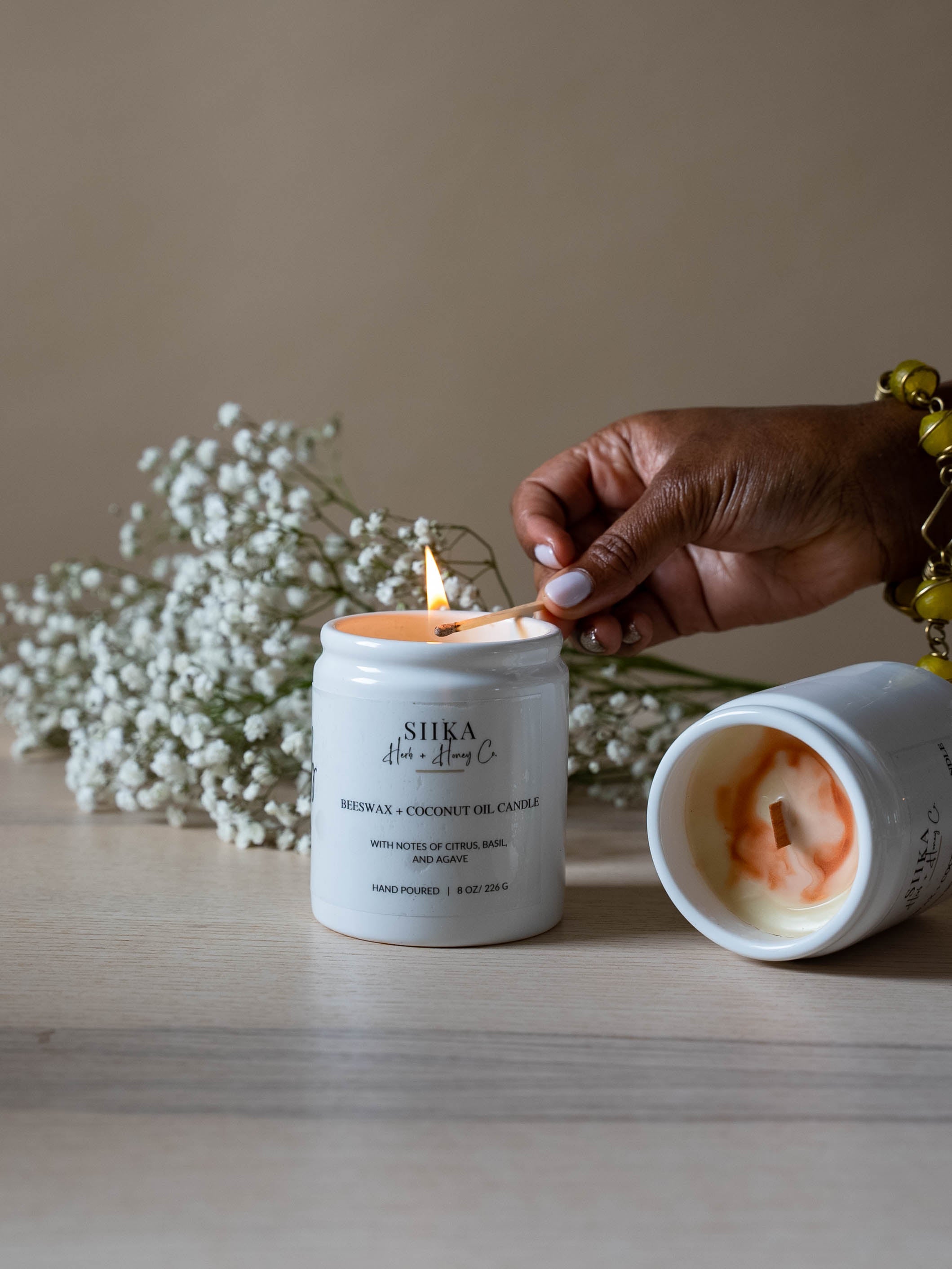 Hand lighting Citrus Basil & Agave beeswax candle with sustainable wood wick, featuring a floral background and minimalist jar design.